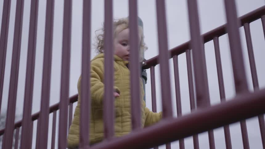 A toddler in a yellow coat walks between railings