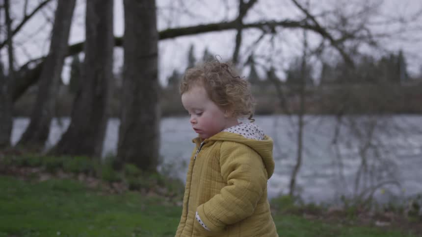A little girl in a yellow coat is walking near a river on an overcast day