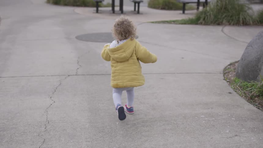 A female toddler in a jacket running down a sidewalk in a park during the day