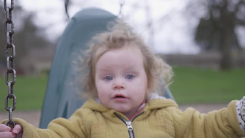 Baby who has blue eyes swings in playground on a cloudy day