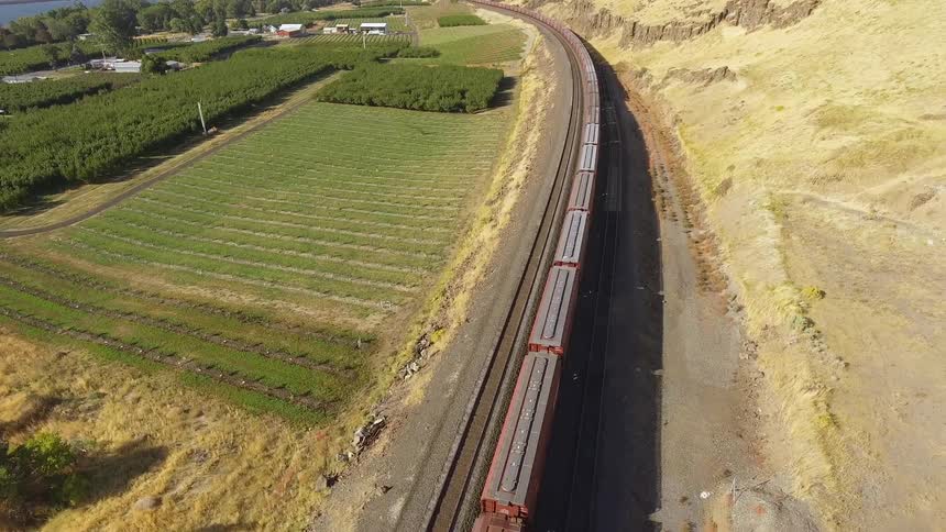 A long train travels past crop fields and orchards