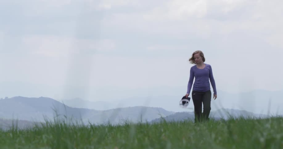 A woman walks through the grass carrying a hat