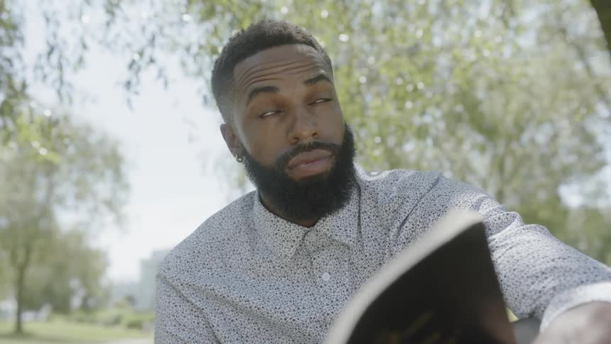 A man is sitting outside reading a book near trees