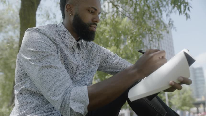 A black man writes in a notebook under a tree near a city center