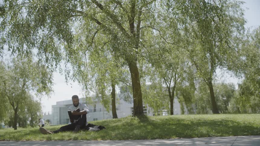 On a very bright and sunny day a man is sitting on the grass under the shade of a tree while he holds a book in his hands
