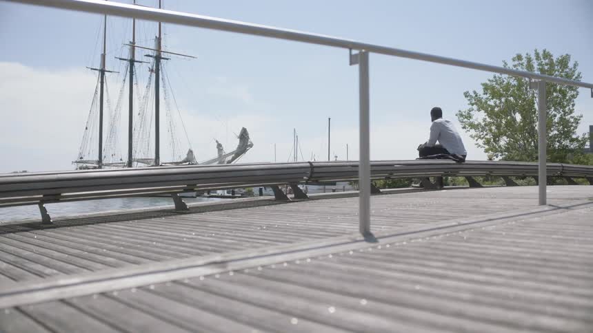 Man sitting on the railing of a bridge looking at a sailboat on the harbor
