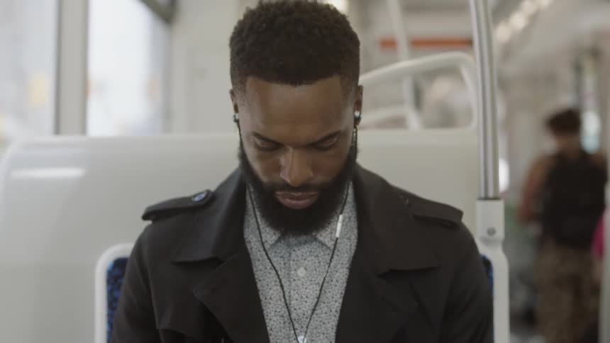 Black man with beard sits on bus with earbuds in ears