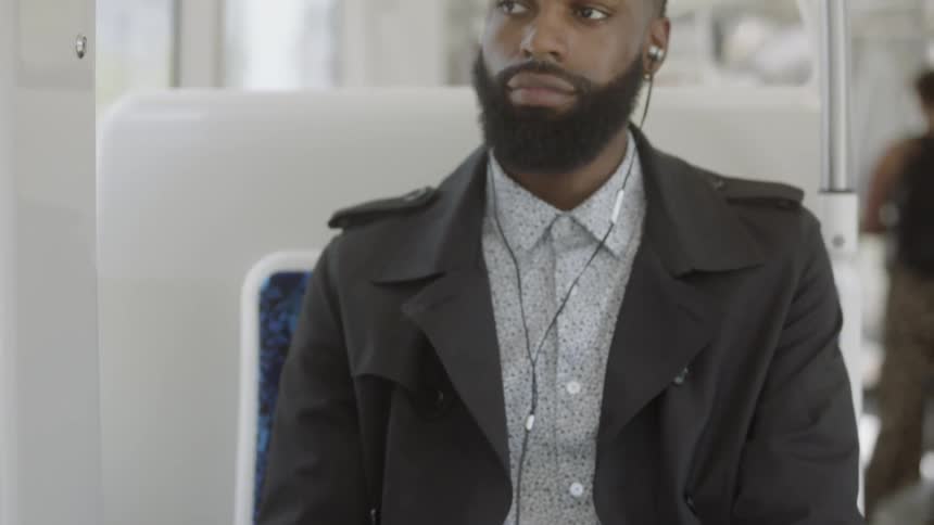 A male wearing a trench coat is sitting on the commuter train while listening to something on his cellphone using earbuds