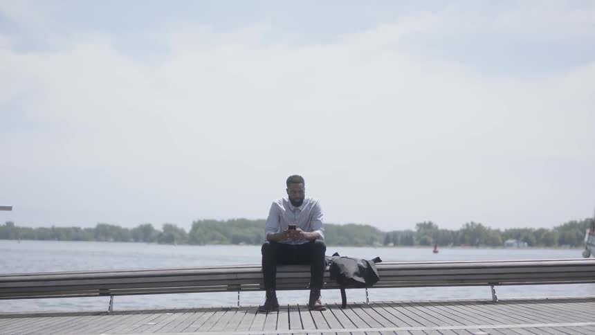 A black male sitting outside on a bench near a river using his phone