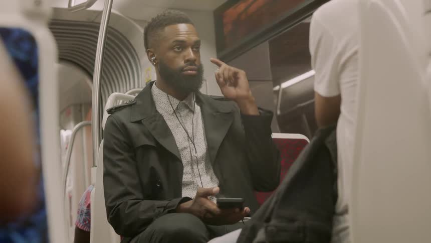 A black man sitting on a bus wearing ear buds using a cell phone in his hand