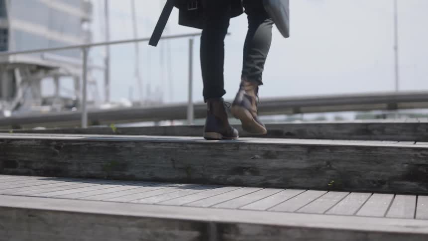 A tall thin man in dark pants and leather boots walks up a number of wooden steps as he carries a coat and a small bag