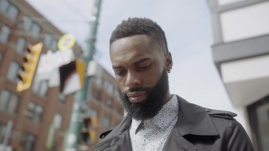 A black man standing outside near buildings looking downward