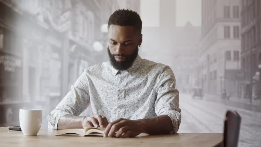 A man reads a book while drinking coffee at a wood table