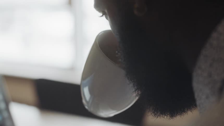 Black man who wears earrings types on laptop and drinks from white mug by day