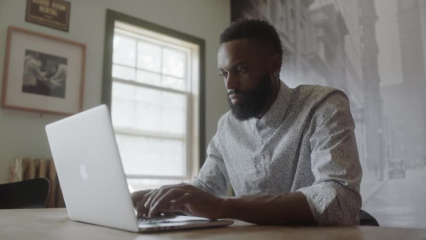 A man sits at a table and types on a laptop then stops to answer a phone call
