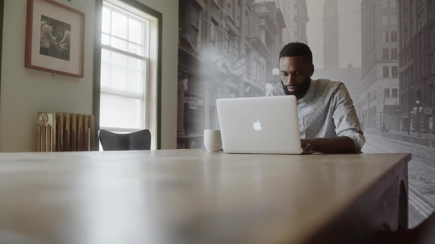 Black man with beard sits and types on laptop in front of photo mural in a room during the day