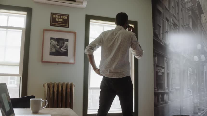 A male who is sitting at a desk in front of a laptop is talking on his cellphone then he stands up and goes over to the window as he is adjusting his shirt and pants while walking