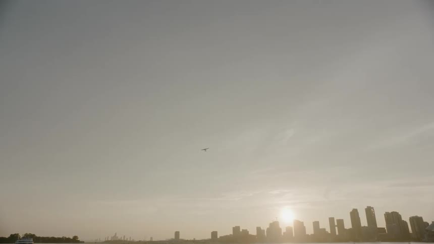 The sun low on the horizon behind a city skyline and reflecting on the water then a plane flying overhead