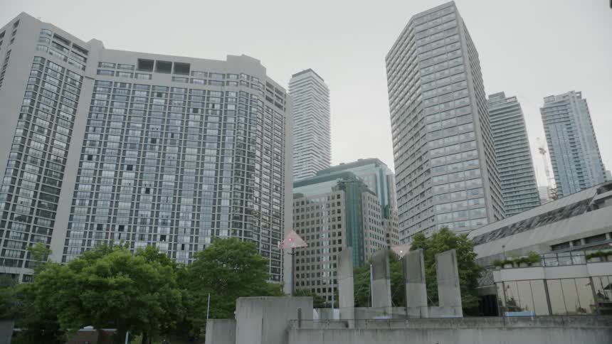 Trees and a cement wall stand in front of several tall buildings in a city skyline on a cloudy day