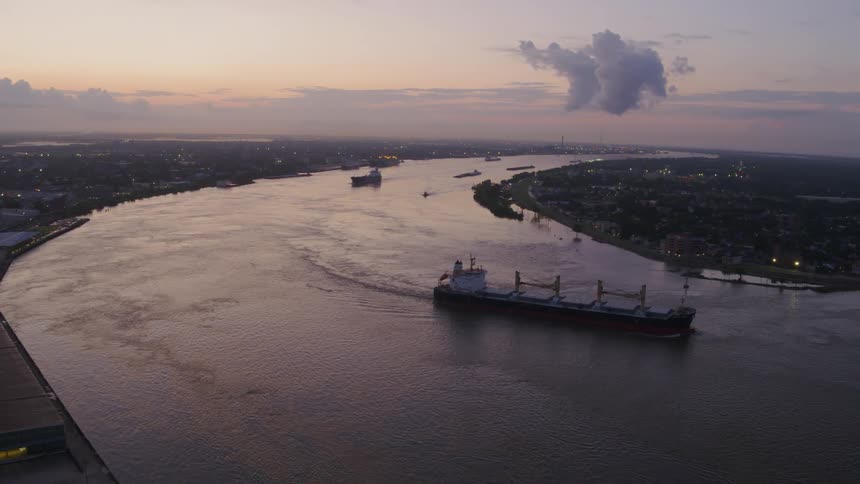 An large shipping vessel with cranes on its deck sails down a river by a city at dusk