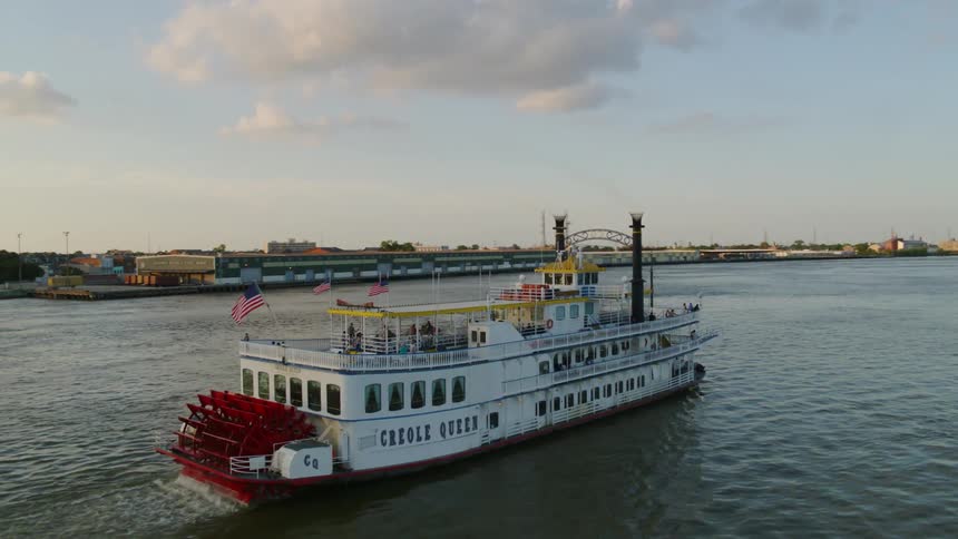 People stand and walk on the upper deck of a large paddle wheel riverboat moving through the water close to shore