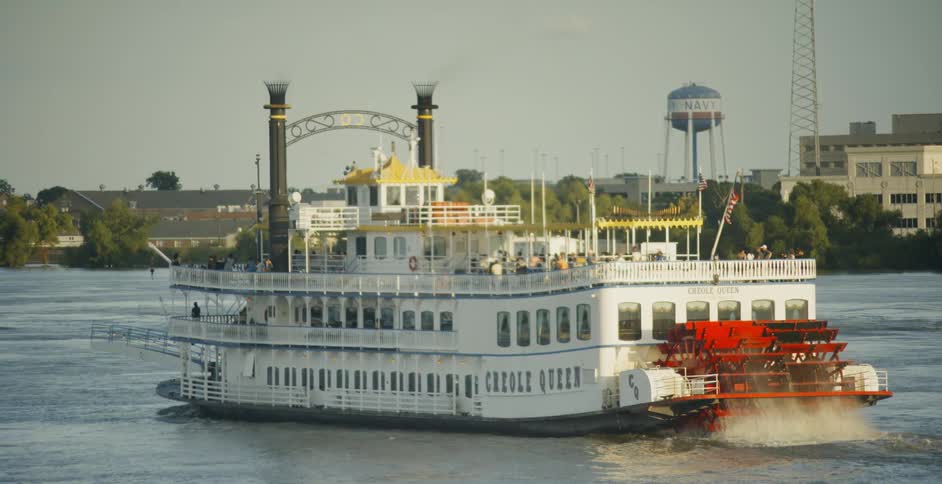 A riverboat with a red wheel turning in the water with trees buildings and a water tower on the other side of the river