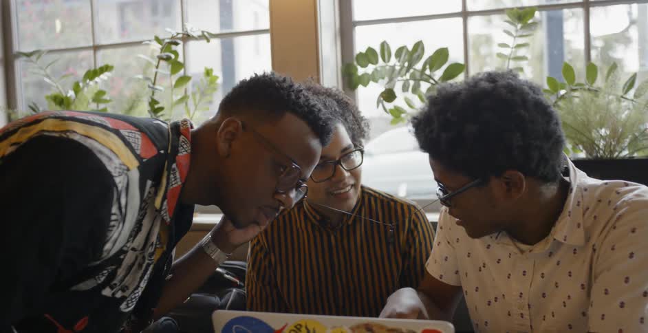 Three young men talk and listen to music on a laptop with headphones they are sharing