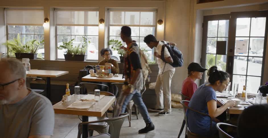Several people are seen sitting around at tables in a coffee shop as two young men with backpacks enter and sit down with their friend