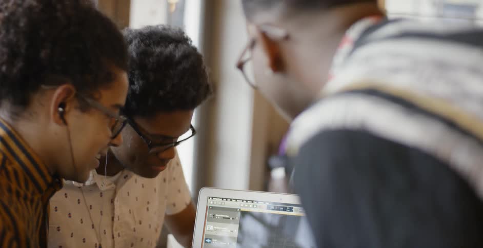 Three men talk and laugh while looking at a laptop