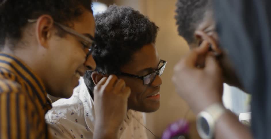 Three men are sitting together listening to music and looking at a computer screen while laughing and talking and moving to the music