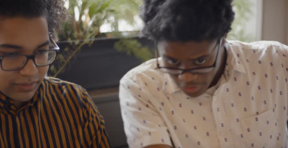 A teenage boy points at a laptop screen and talks to his friend sitting beside him