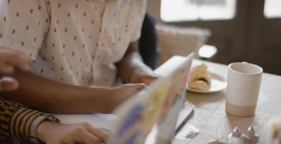 A young map using a laptop sitting next to a young man writing in a notebook at a dining table