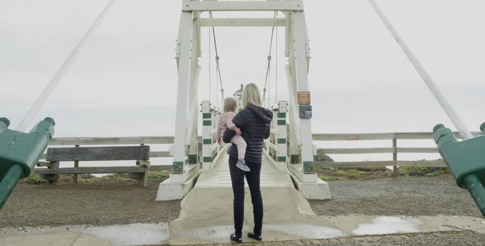 A woman with very light blonde hair stands holding a toddler in front of a suspension bridge on an cool overcast day