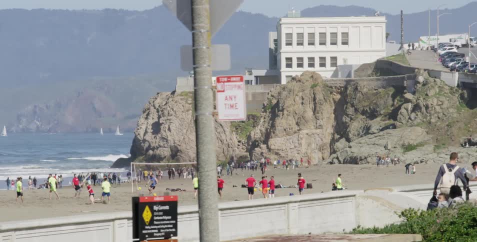 People play beach soccer at the beach while waves hit the shore with distant mountains on a sunny day