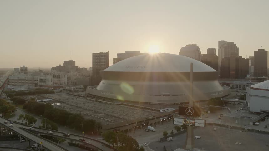 The sun sets behind a large sports stadium with skyscrapers in the background