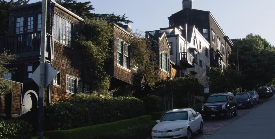 A street on the side of a hill is lined with several very lovely residences and cars are parked alongside the street as well