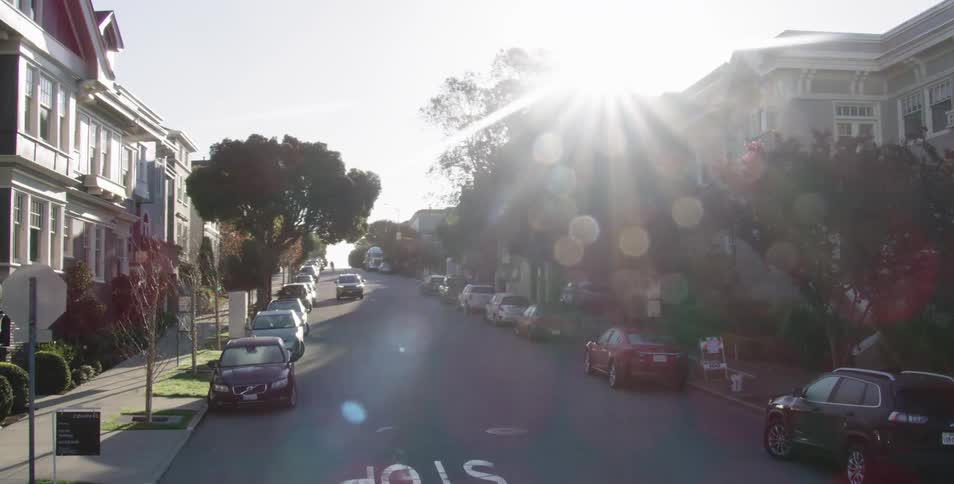 A road lined with many nice residential building runs up a hill and many parked cars line this street on a bright and sunny day