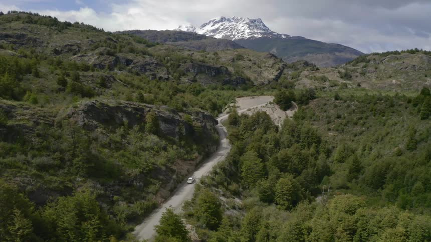 A narrow road cuts through an expansive forest at the base of a snow capped mountain