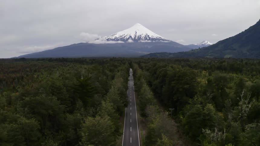 A paved two lane road cuts a straight line through a dense forest that lies below a towering snow capped mountain