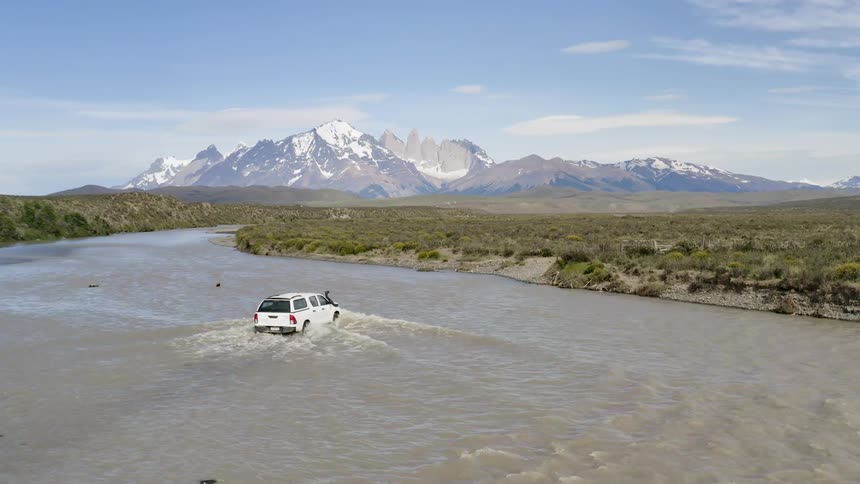 A truck with a covered bed drives through a river with mountains in the background