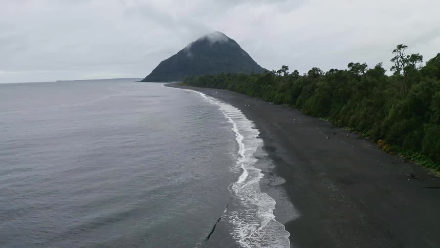 Waves crash onto the beach front of a forest island with a volcano in the background during a cloudy day