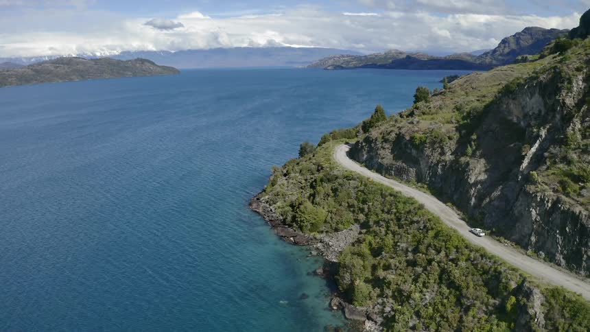 A white car drives along a mountain road next to the ocean