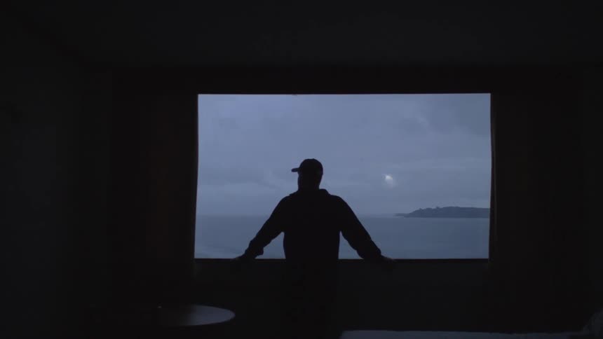 A man stands in front of a large window looking out over a body of water