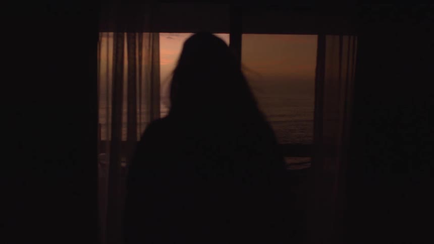 A silhouette of a woman in a dark room walking toward a window looking at the sun setting over the ocean