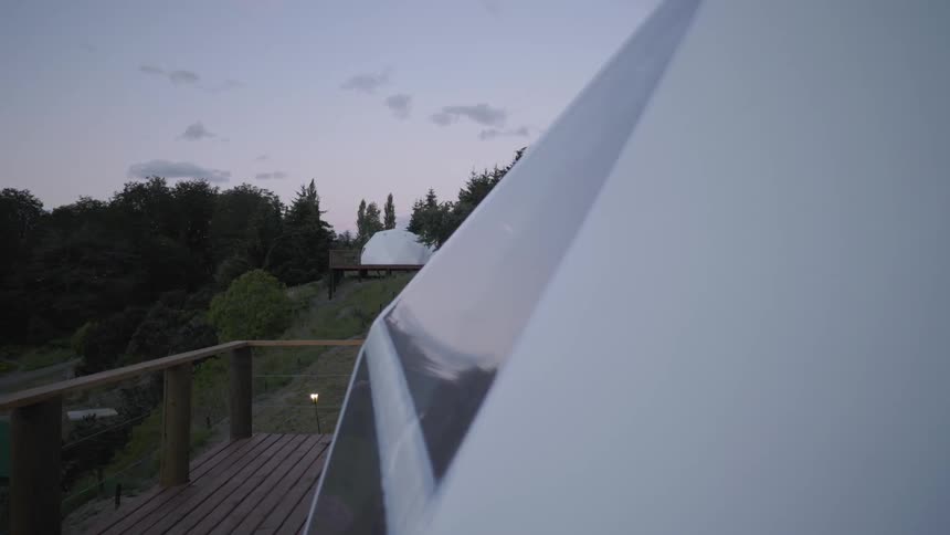 A deck to a building at dusk in the woods with a similar building in the distance
