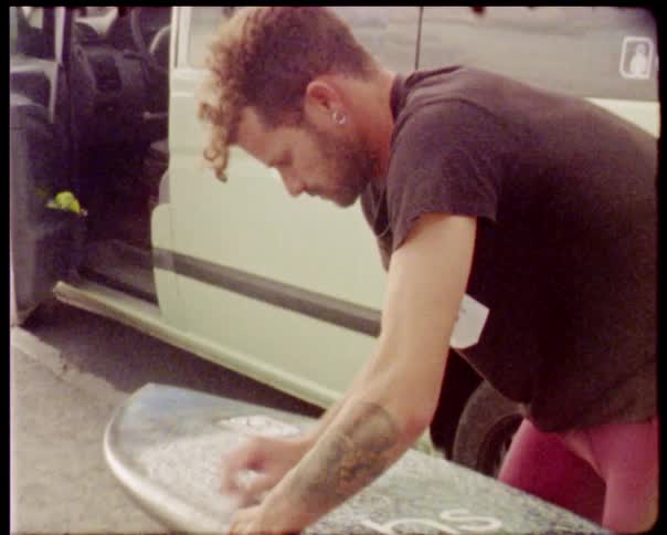 A man wearing red shorts who has a tattoo on his left arm is standing outside of his white van in a parking lot while waxing a surfboard
