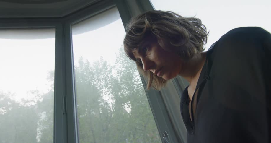 A woman stands in a room full of windows in a house on a cloudy day in the city