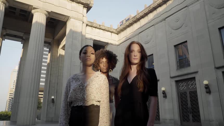 Three women stand partially illuminated by a low sun next to classical pillars