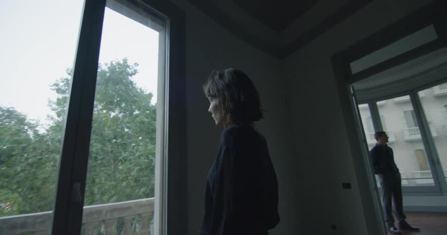 A woman stands at a window looking out longingly as a man stands in the next room