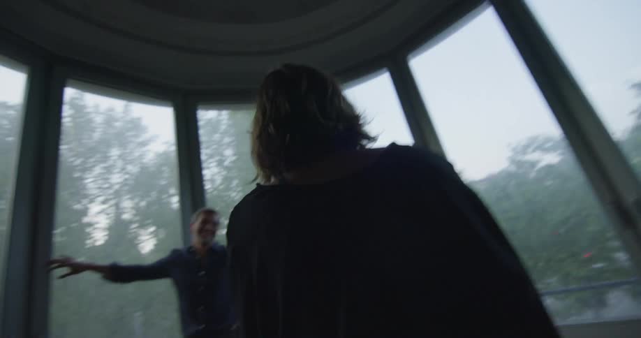 A woman walks into a room with a circular wall of picture windows toward a man leaning on one of the windows